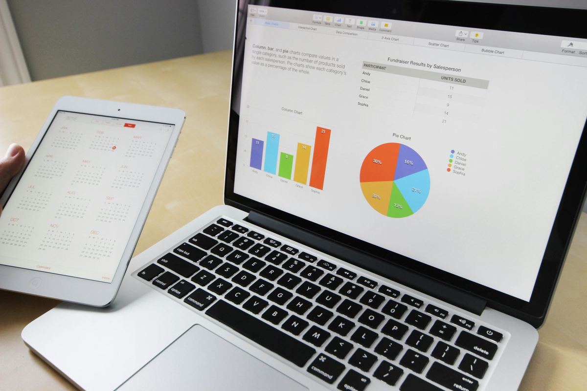 A hand holding a tablet showing a calendar beside a laptop displaying a sales dashboard with bar and pie charts.